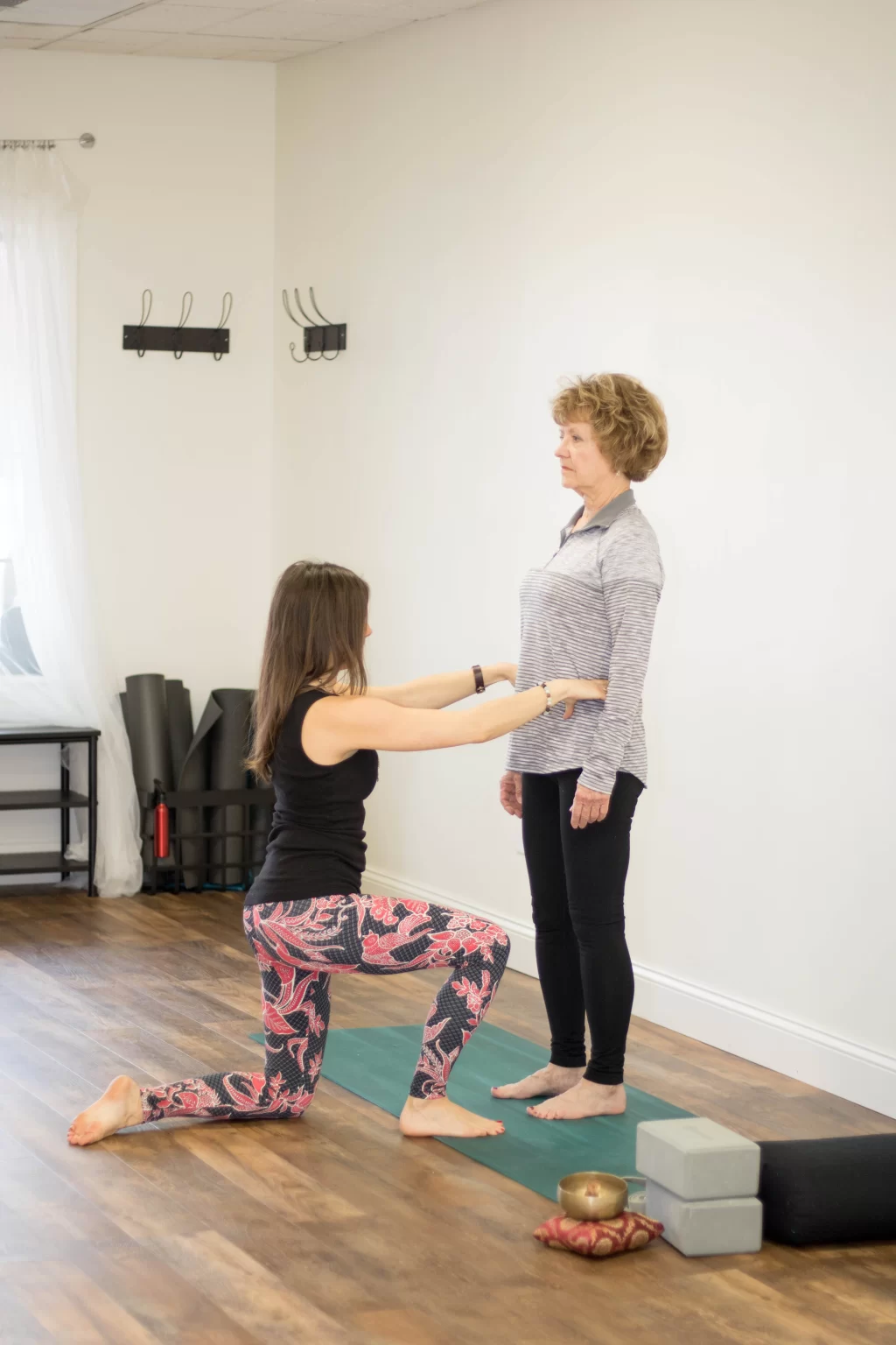 Woman kneeling with arms extended in front of another woman standing, both on a yoga mat in a room with wooden floor and exercise equipment.
