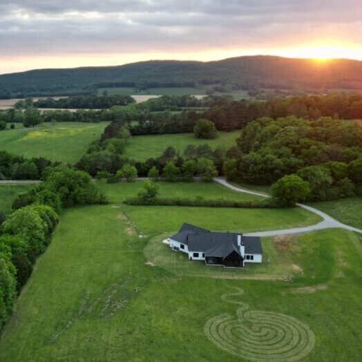 Drone image of rolling lush green hills, a house, and a labyrinth with many surrounding trees and a sunset.